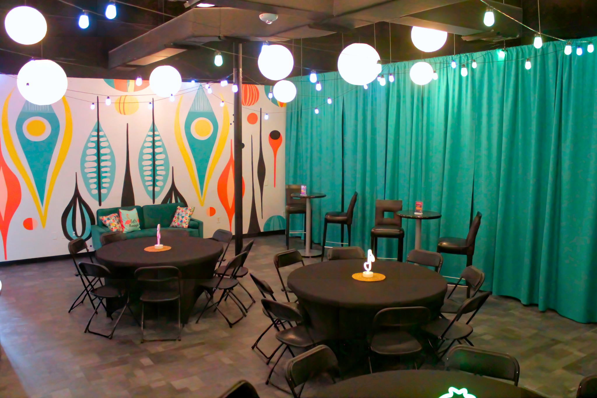 Private party room with round tables, black linens, a teal sofa, a colorful mural, and glowing globe lights