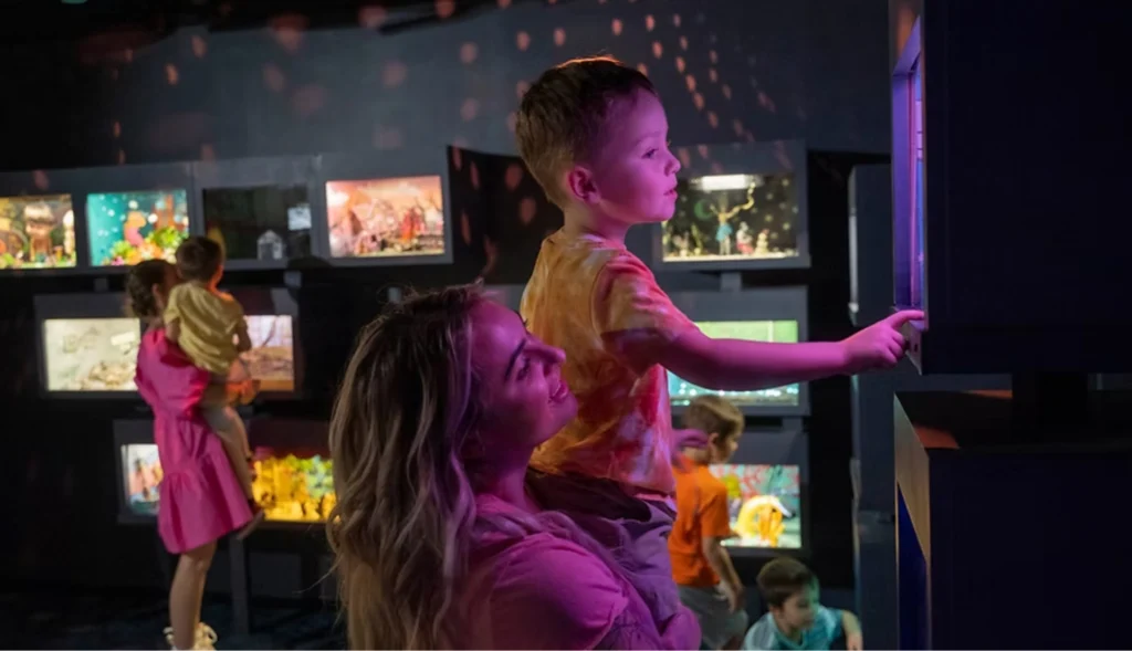A woman holds a child as they interact with a glowing diorama box in a room filled with immersive art exhibits