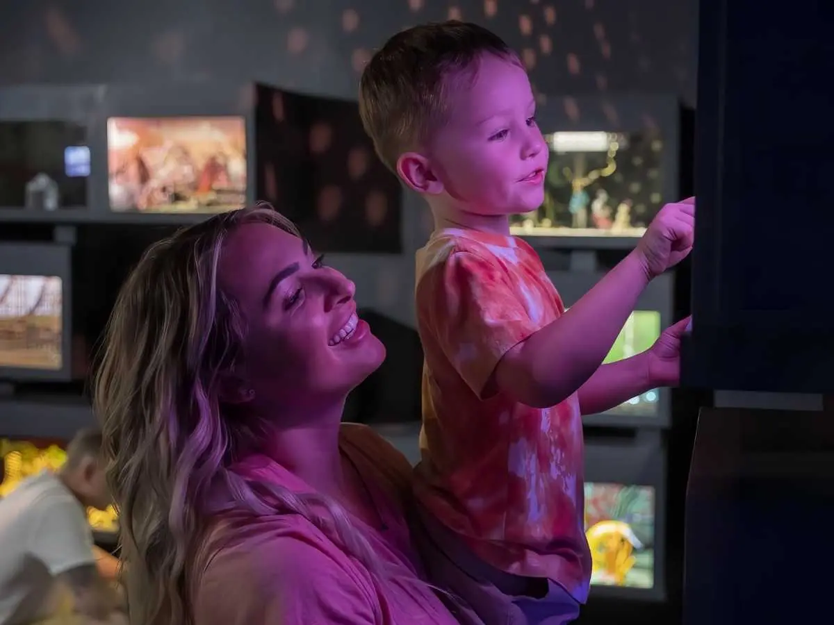 A woman holds a young boy as he interacts with a glowing art installation in an immersive gallery