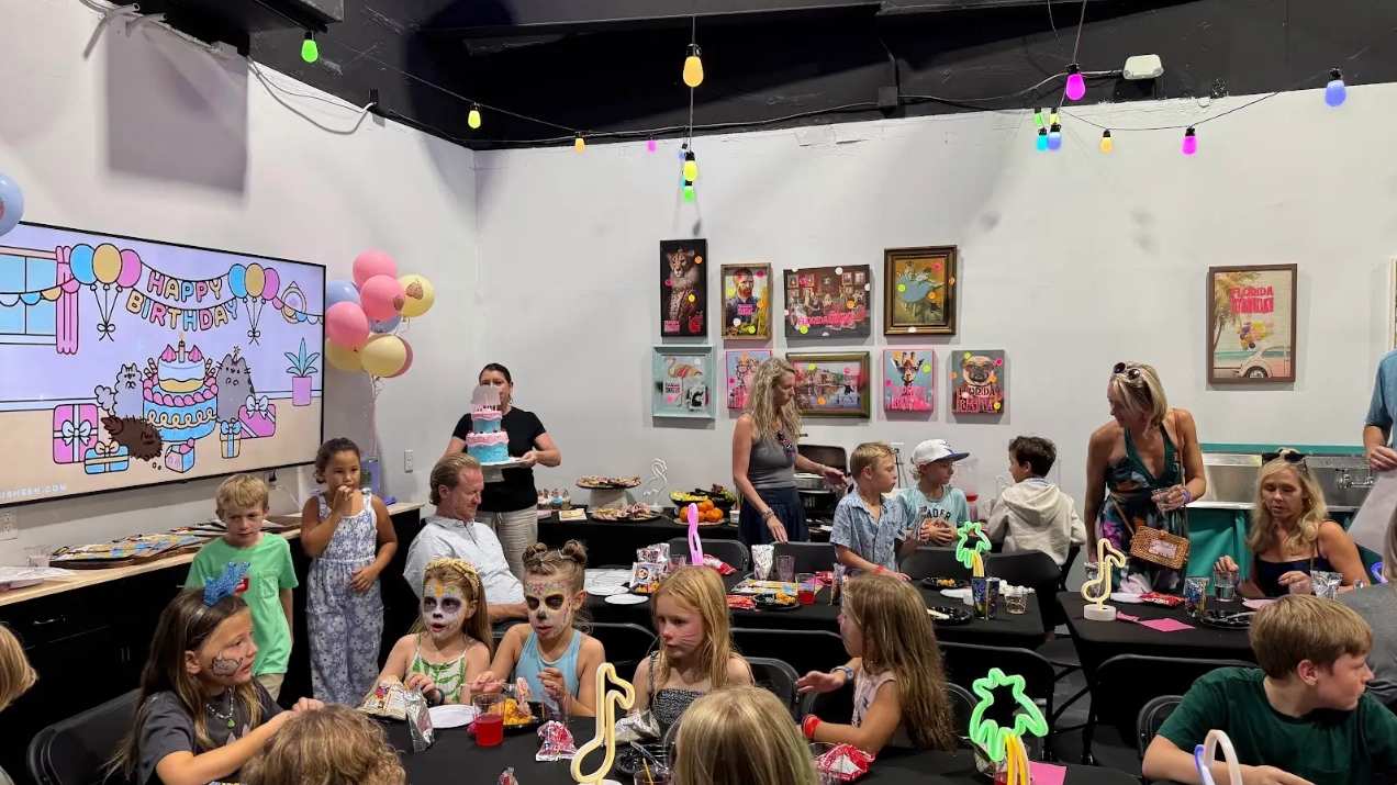Children with face paint and adults celebrate a birthday in a colorful party room with art and a tiered cake
