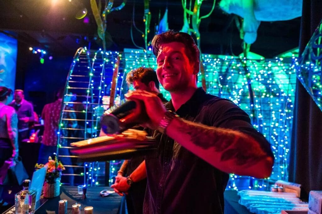 Smiling bartender at a wedding shaking a cocktail in front of vibrant, glowing blue and green light installations at FloridaRAMA